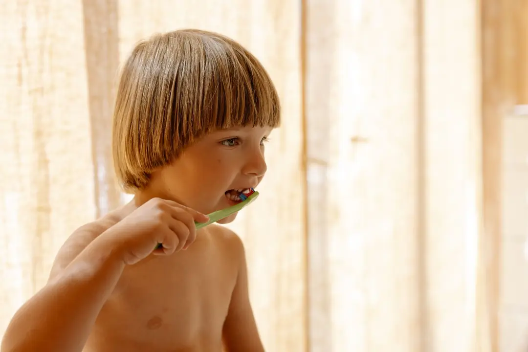 A young boy brushing his teeth in soft natural light, holding the brush in his own fist
