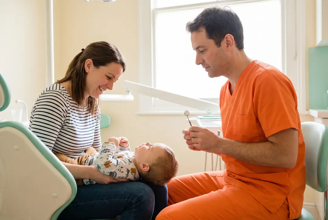 A dentist and a parent sitting knee to knee with a toddler lying back across both their laps, the child looking up at the parent's face