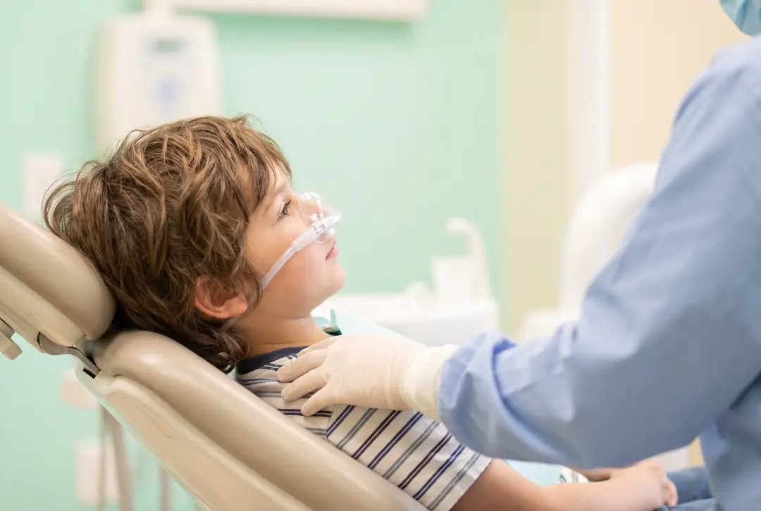 A child lying back in a dental chair with a small nasal hood for nitrous oxide in place, watching a ceiling-mounted screen while a dentist sits alongside